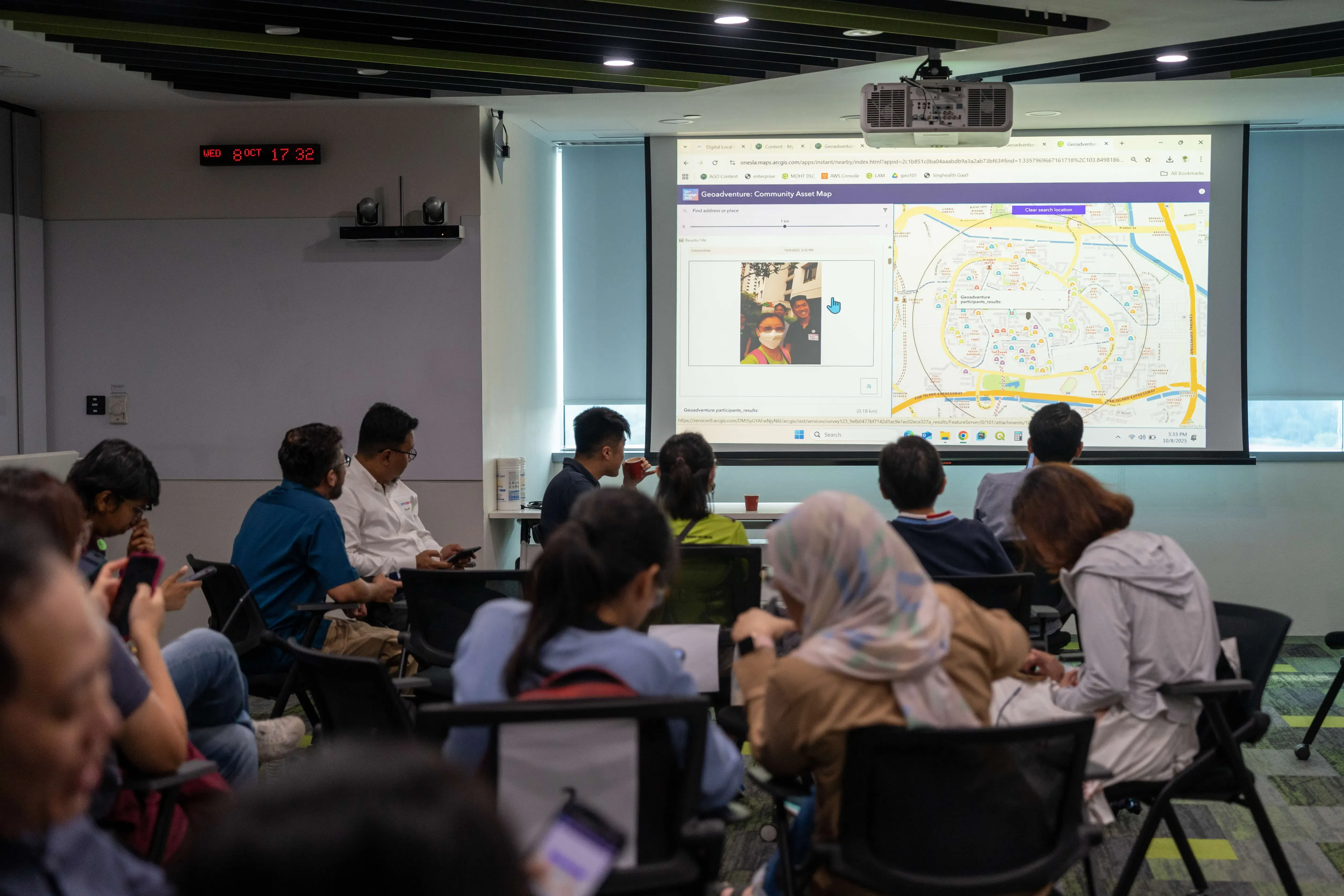 A group of people seated in a conference room facing a projected screen showing a community map application.